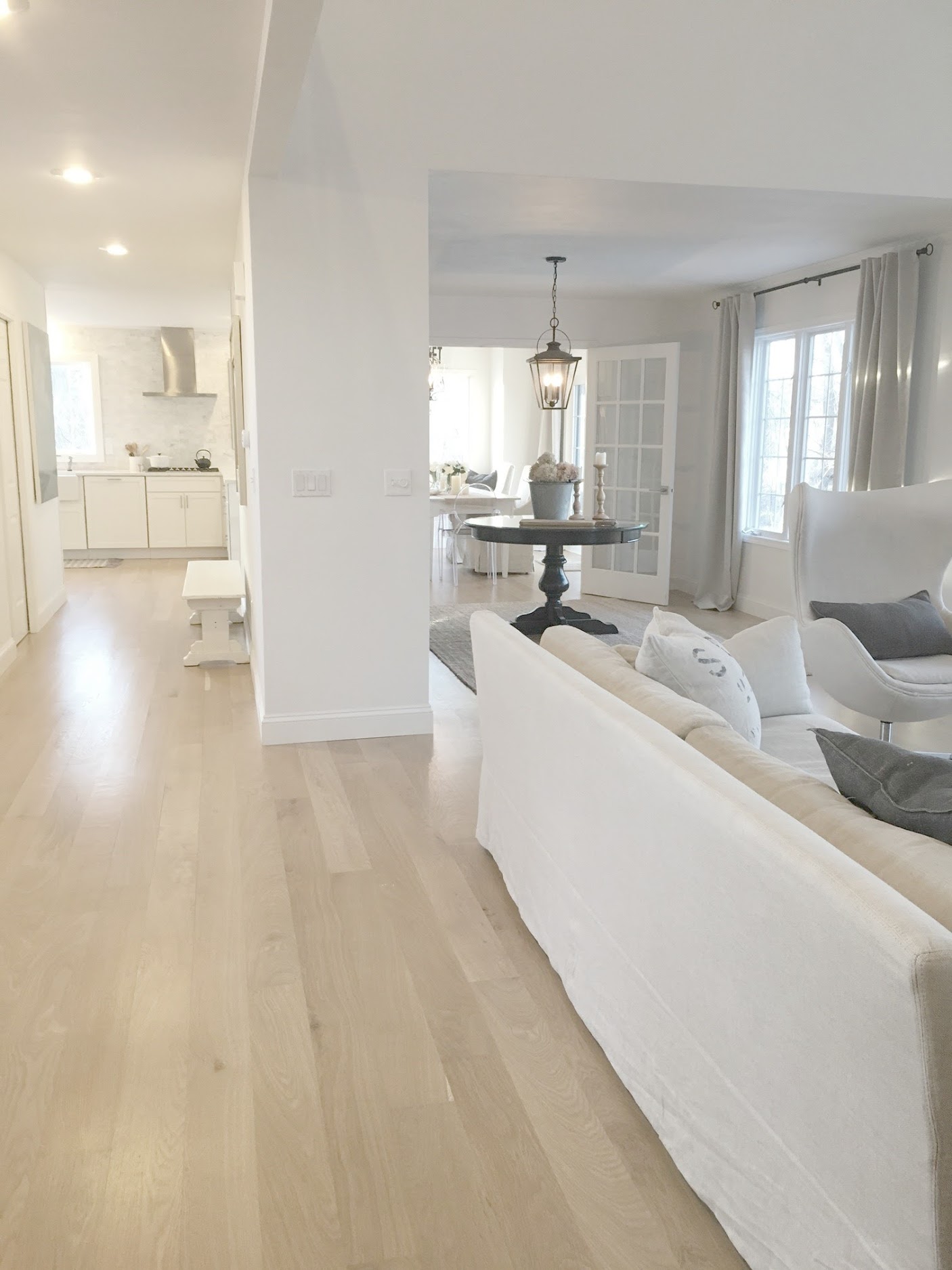 Serene white living room with white oak flooring, Belgian linen upholstery and spare decor in a rustic elegant cottage with open floor plan. Hello Lovely Studio is a blog with French country inspired interiors, French farmhouse decor, and interior design inspiration overflowing with timeless and tranquil charm.