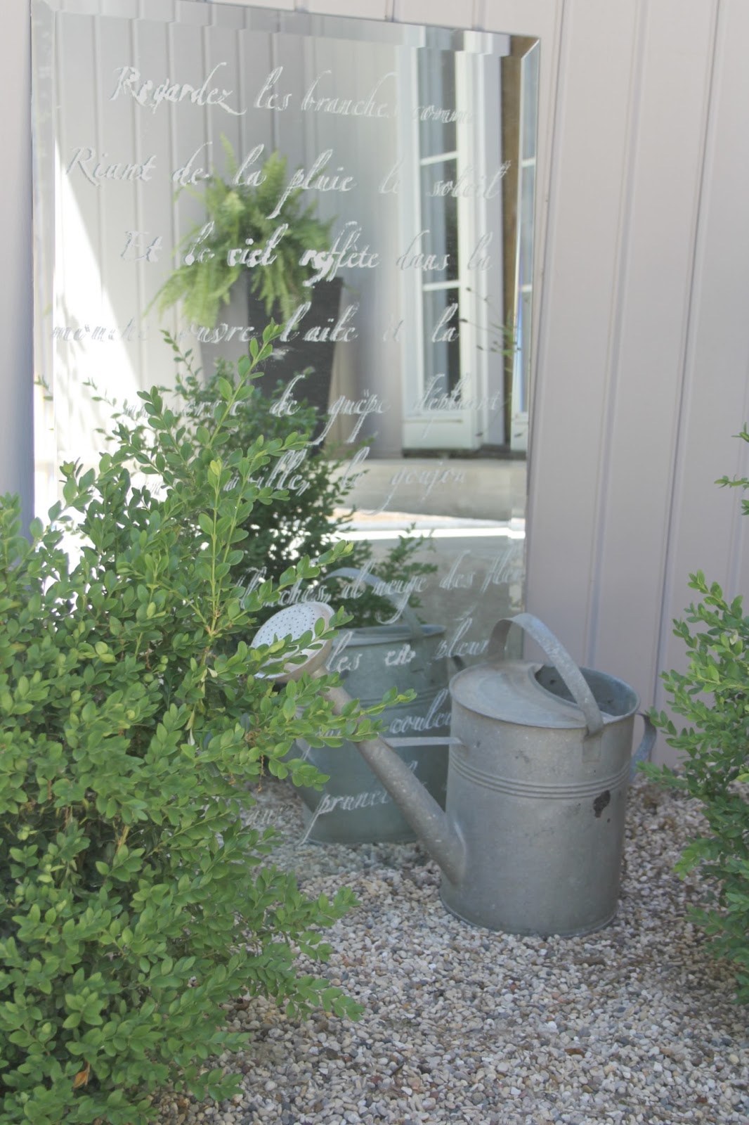 French farmhouse decor style in a courtyard garden with pea gravel, a vintage watering can, and a mirror stenciled with a French poem. #hellolovelystudio #frenchcountry #frenchfarmhouse #courtyard #stenciled #mirror